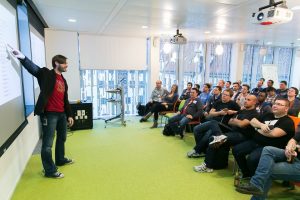 Dirk Primbs on stage with one arm raised, addressing a seated audience in a room with a green floor.